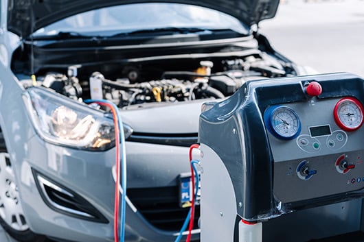 Car undergoing an AC recharge service with the hood open and AC maintenance equipment connected.