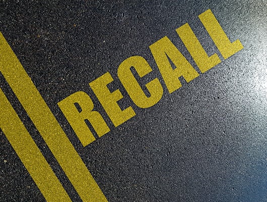 The word "RECALL" is painted in bold yellow letters on a dark asphalt road, with two yellow lines running parallel to the text. This image symbolizes the concept of vehicle recalls and the importance of addressing them.